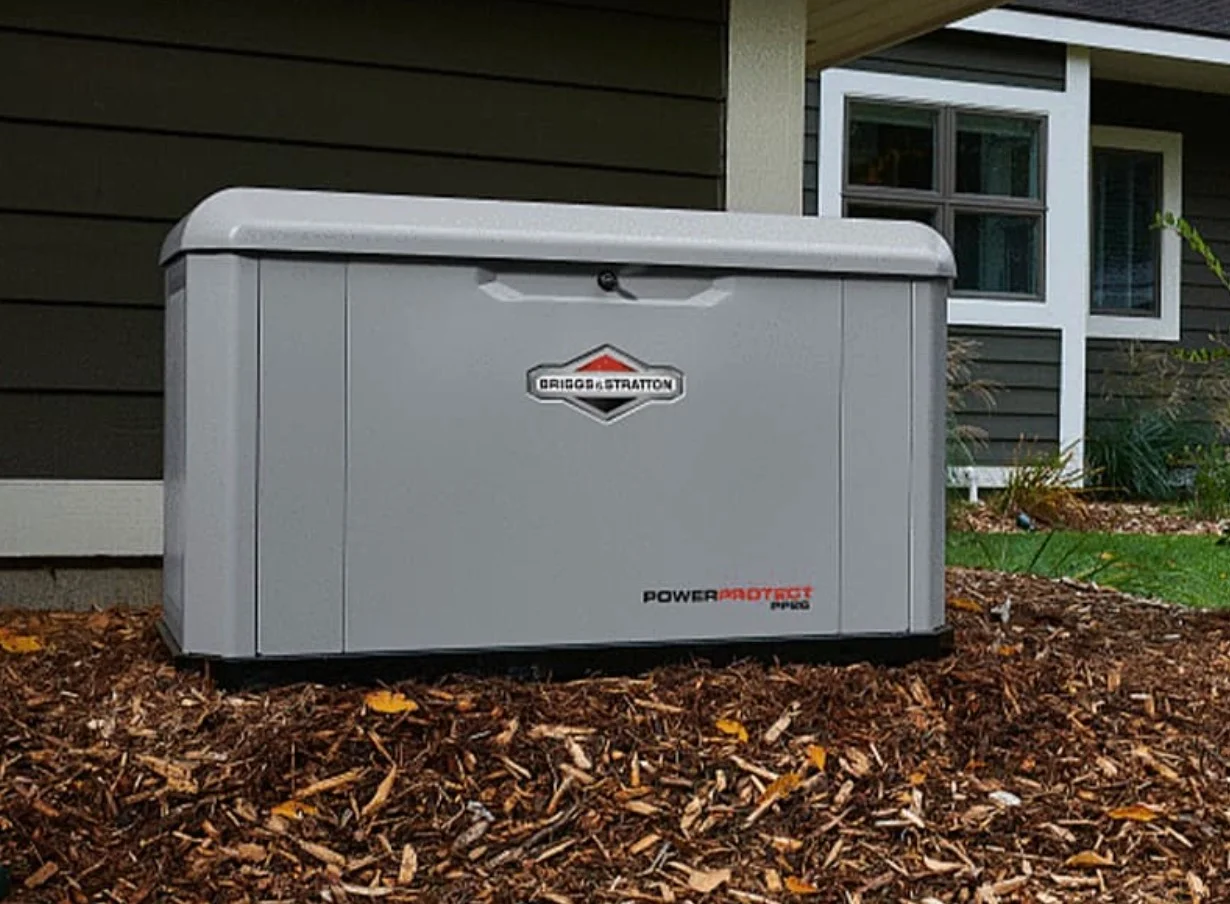 A gray Briggs & Stratton generator sits on mulch beside a house.